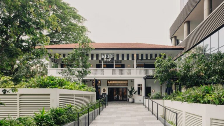 Two-story white café with a striped awning, balcony, and lush plants along a paved walkway.