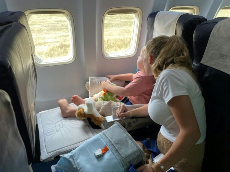 Mother and young child seated by a plane window; the child plays with a toy, a stuffed dog rests on the footrest nearby.