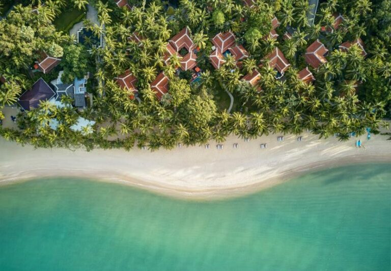 Aerial view of a tropical beach with a line of palm trees and red-roof villas along the shore, turquoise water lapping the white sand, viewed from above