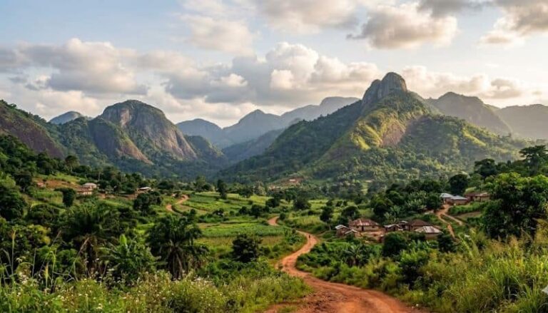 Dirt road winding through a green valley with small houses and lush hills, mountains in the distance under a cloudy sky.