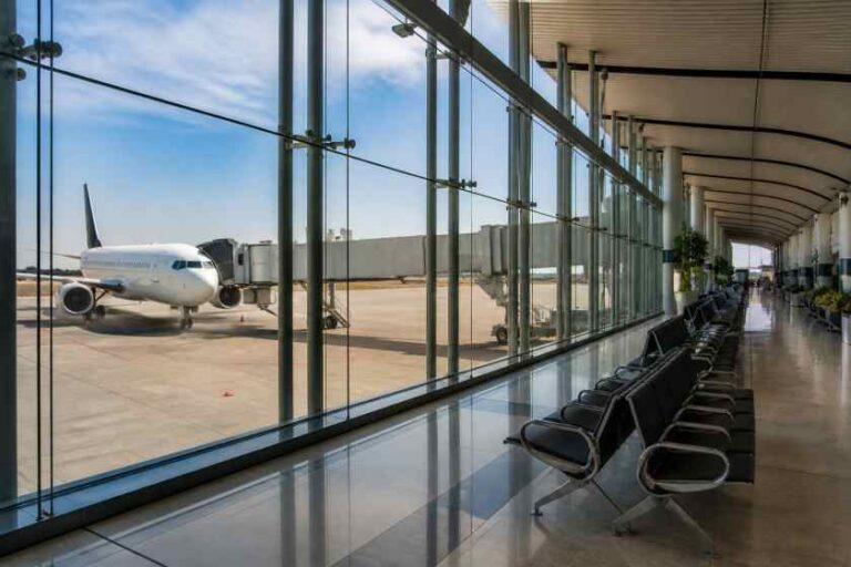 Airplane parked at a gate viewed through a glass-walled airport terminal, with row of black chairs along the corridor outside the window.