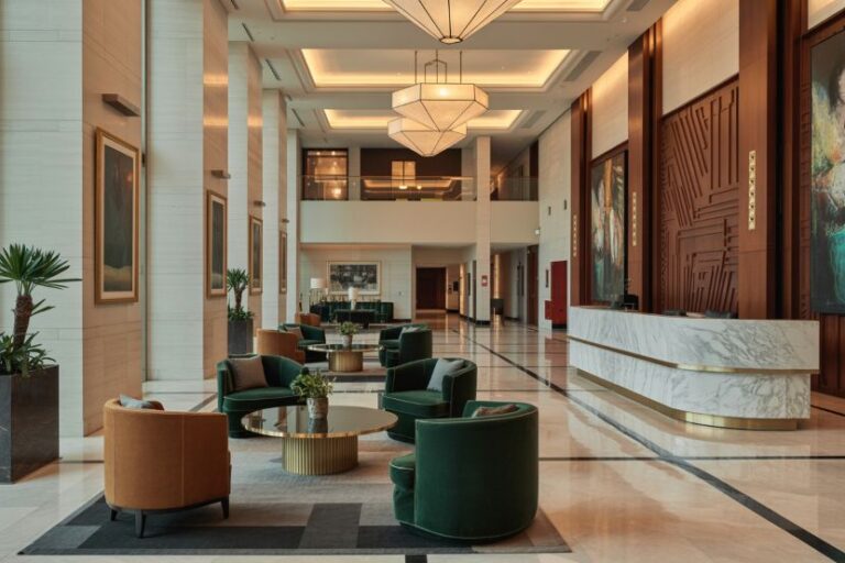 Hotel lobby with green and tan armchairs around round glass tables, marble floors, tall columns, and a curved marble reception desk under geometric chandeliers.