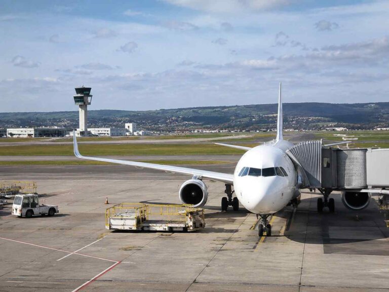 White passenger jet connected to a jet bridge at a gate on a tarmac, with a distant control tower and runway in the background