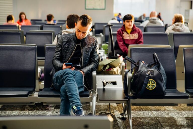Man in a black leather jacket sits in a busy airport waiting area, looking at his phone with a backpack and bag beside him.