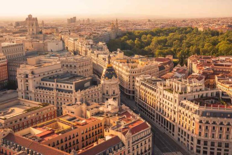 Aerial view of a sunlit European city center with ornate cream buildings and a large green park in the distance, at sunset credibility for a historic district.