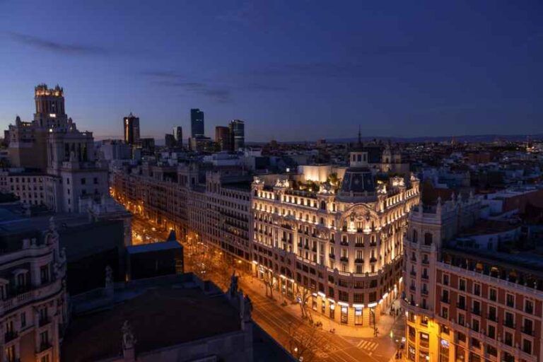 Twilight cityscape with ornate, illuminated buildings along a broad street, seen from an elevated vantage point.