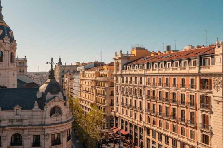 A European city street with ornate beige buildings, balconies, and café terraces under a clear blue sky.