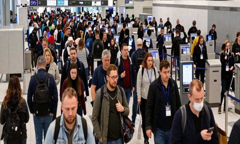 Crowd of travelers in a busy airport check-in area, lined up at self-service kiosks.