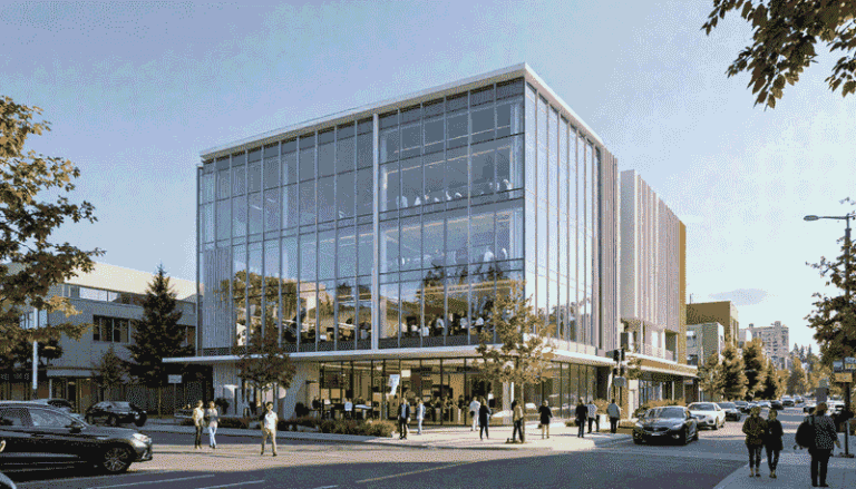 Modern glass-fronted building with people walking and cars on a sunny street outside.
