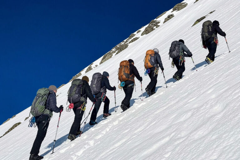 Group of hikers with large backpacks climbing a snowy slope under a clear blue sky.
