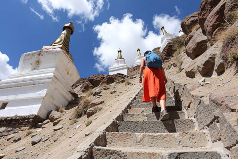 A hiker in an orange dress with a blue backpack climbs stone steps along a rocky hillside beside white Buddhist stupas under a blue sky.