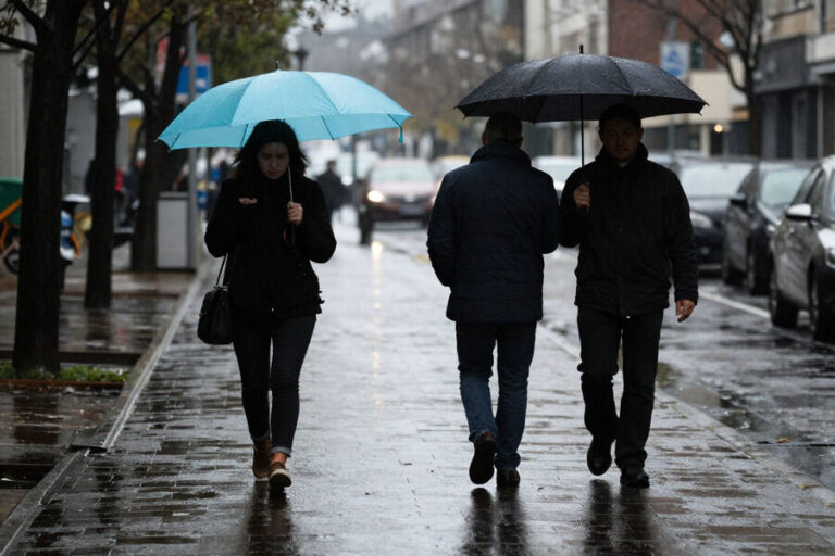 People walking on a rainy city sidewalk holding umbrellas, the turquoise umbrella prominent on the left side.