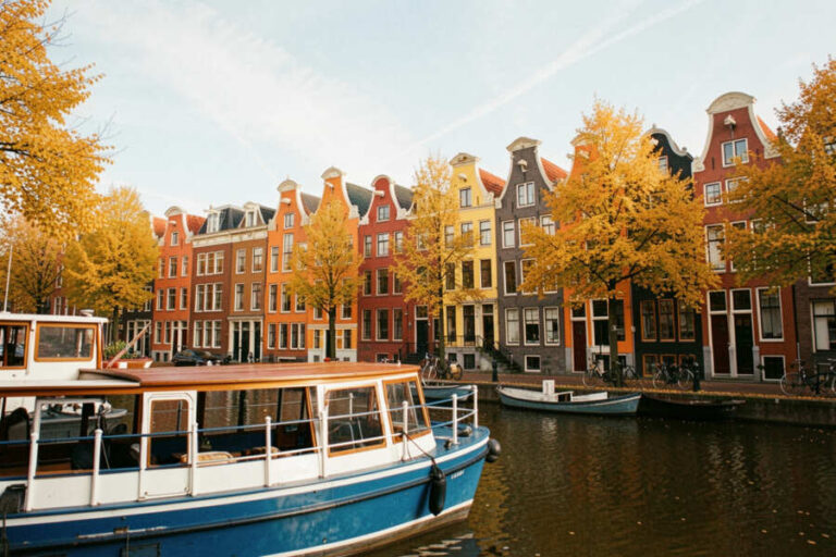 Row of colorful Dutch houses along a calm canal, autumn trees in orange and yellow, with a blue boat in the foreground.