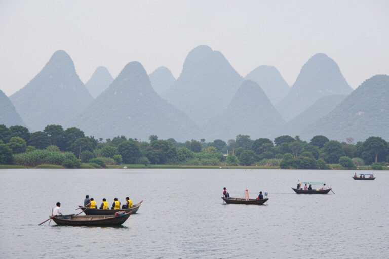 Boats with people wearing yellow life jackets on a calm lake with green shoreline and karst hills in the distance