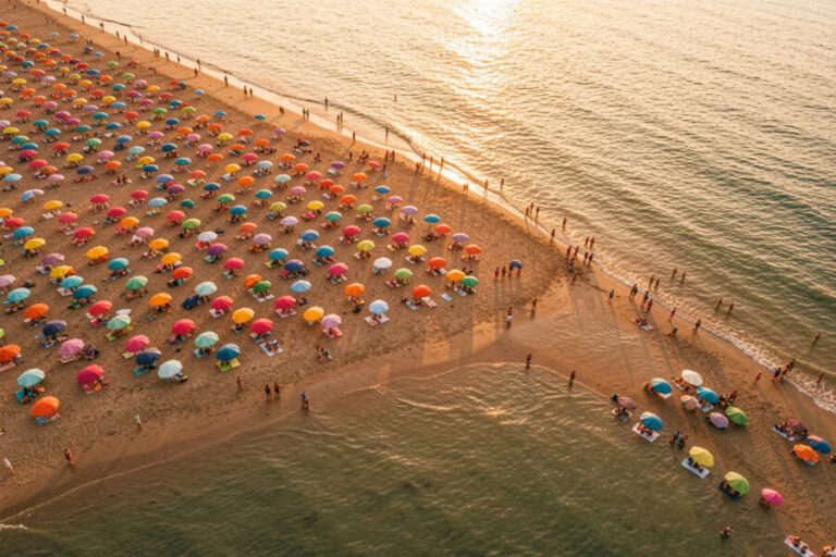 Crowded sandy beach with dozens of colorful umbrellas arranged in rows along the shore.