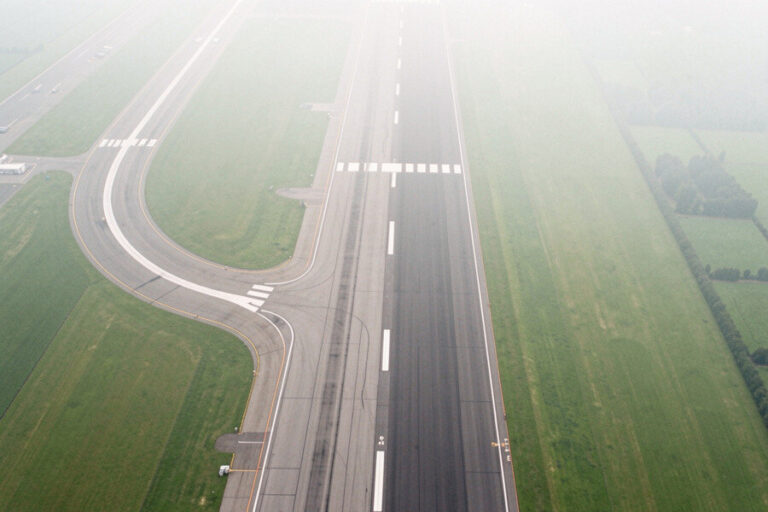 Aerial view of a runway with parallel taxiways and a curved taxiway, green fields on both sides, lightly obscured by fog.