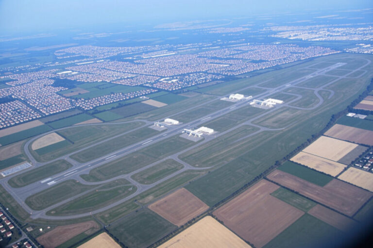 Aerial view of a large airport with parallel runways, taxiways, and white terminal buildings, surrounded by fields and a grid of houses nearby.