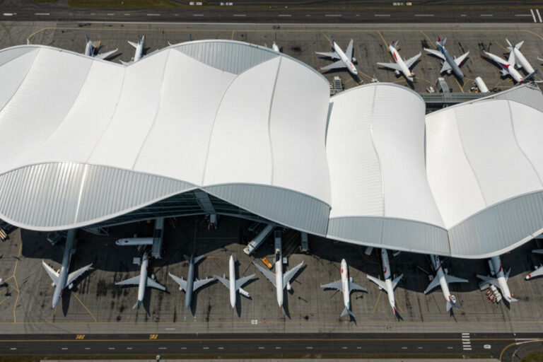 Aerial view of an airport terminal with a white wavy roof and numerous parked airplanes on the tarmac at gates.