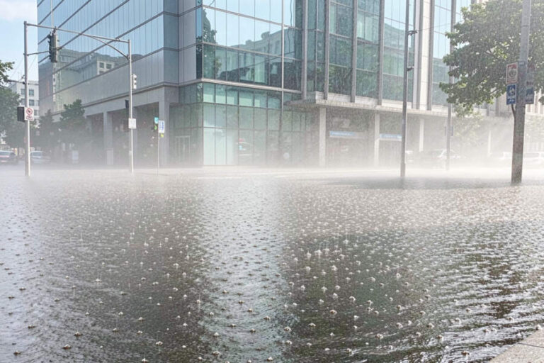 Flooded city street in front of a glass office building, water rising and misty air after heavy rain.
