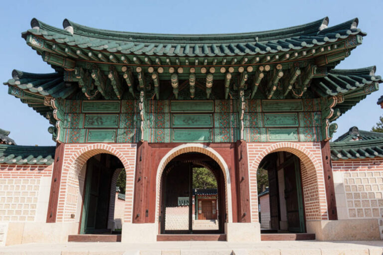 Traditional Korean gate with green tiled roof and ornate wooden carvings, brick arches, and mirrored courtyard beyond.