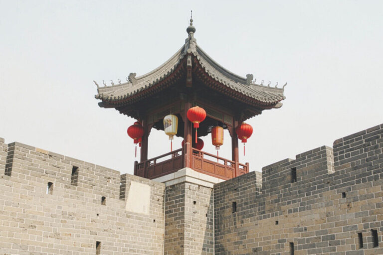 Traditional Chinese pavilion with red lanterns atop a historic stone wall/fortress.