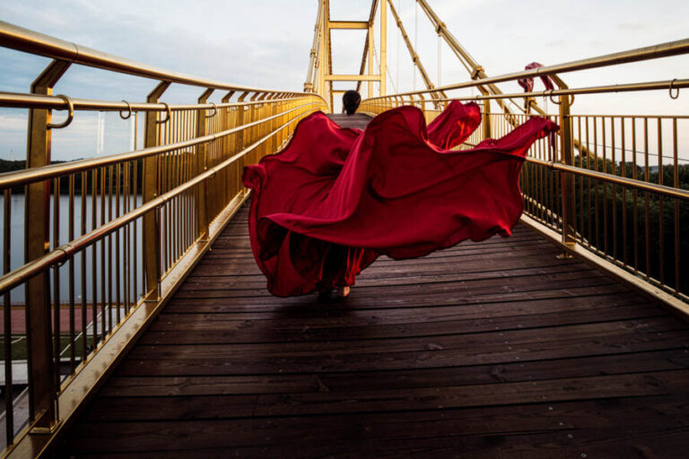 Person in a flowing red cape walking fast on a golden pedestrian bridge, cape billowing behind them