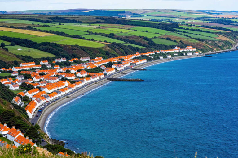 Coastal village with white houses and red roofs along a curved blue bay, green farmland beyond.