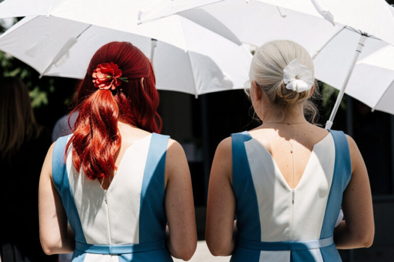Two women in matching blue and white dresses stand with backs to the camera under white umbrellas; one has red hair with a flower, the other blonde with a white flower in her hair, at an outdoor event.