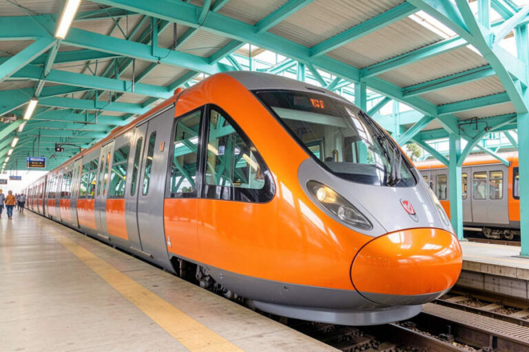 Front view of an orange and gray passenger train at a station with turquoise metal beams overhead.