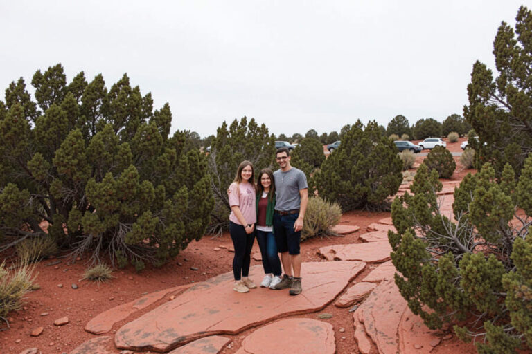 Three people pose together on a red rock path in a desert, with green shrubs and parked cars in the distance.