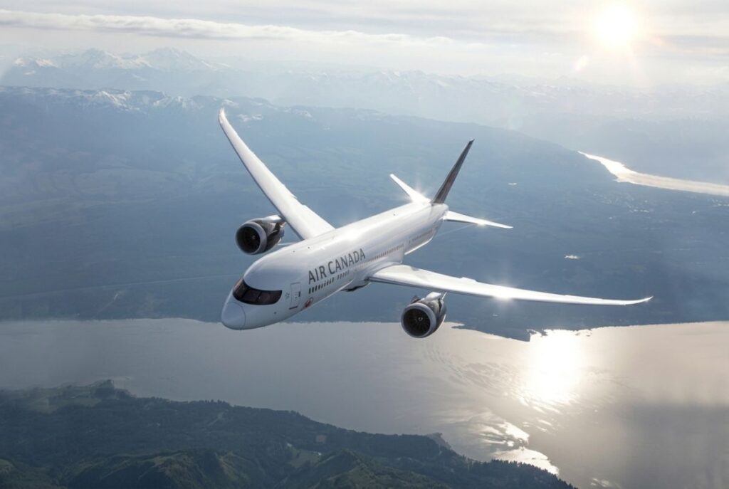 Air Canada passenger jet flying over a mountainous coastline with sun glare on the water below.