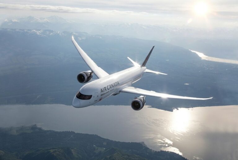 Air Canada passenger jet flying over a mountainous coastline with sun glare on the water below.