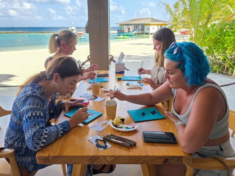 Four women sit at a wooden beachside table painting small teal canvases, with the sea and boats in the background.