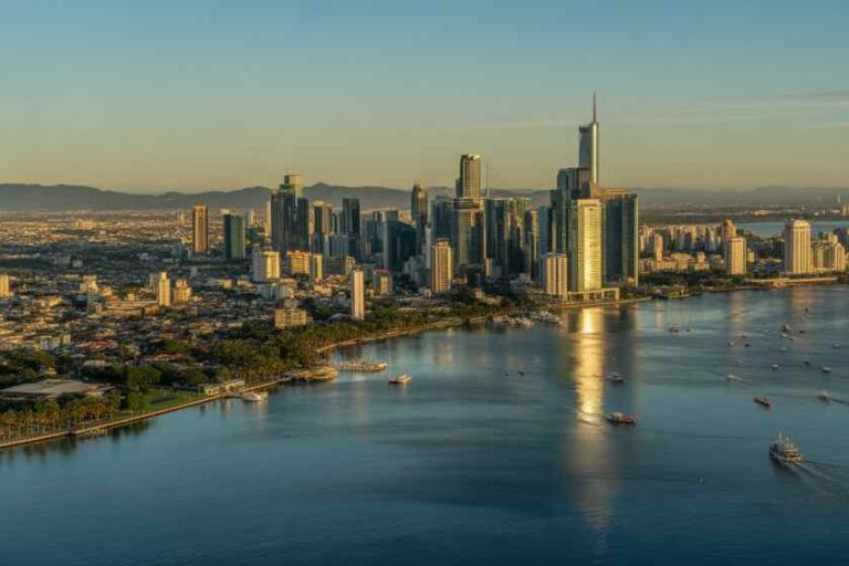 A dense downtown skyline of tall buildings along a wide river at sunset or dusk, with calm water reflecting the city lights.