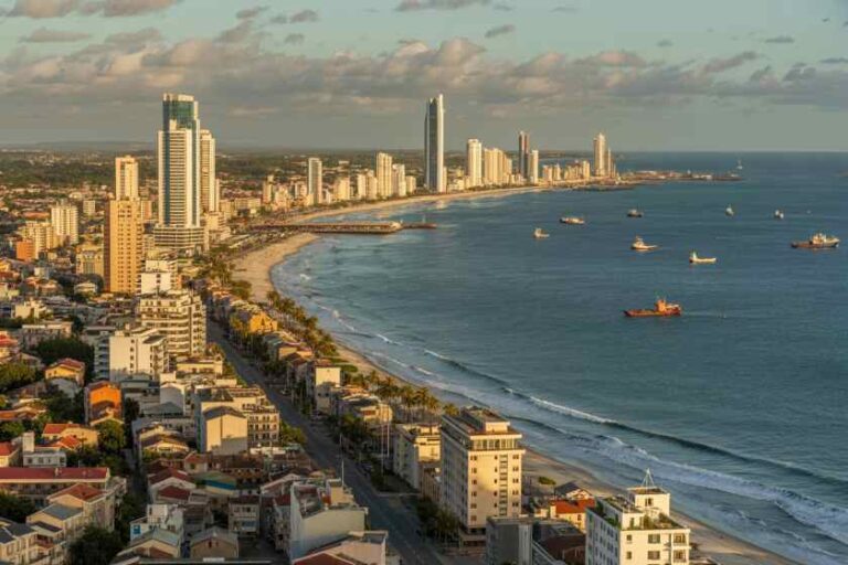 Aerial view of a sunny coastal city with high-rise buildings along the beach and boats in calm ocean water.