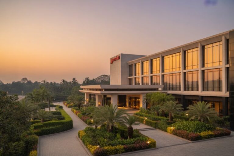 Hotel building at sunset with palm trees and manicured gardens along a curved driveway, inviting entrance visible in warm light.