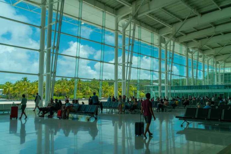 Bright airport terminal with floor-to-ceiling glass walls, travelers seated with luggage waiting by blue chairs.