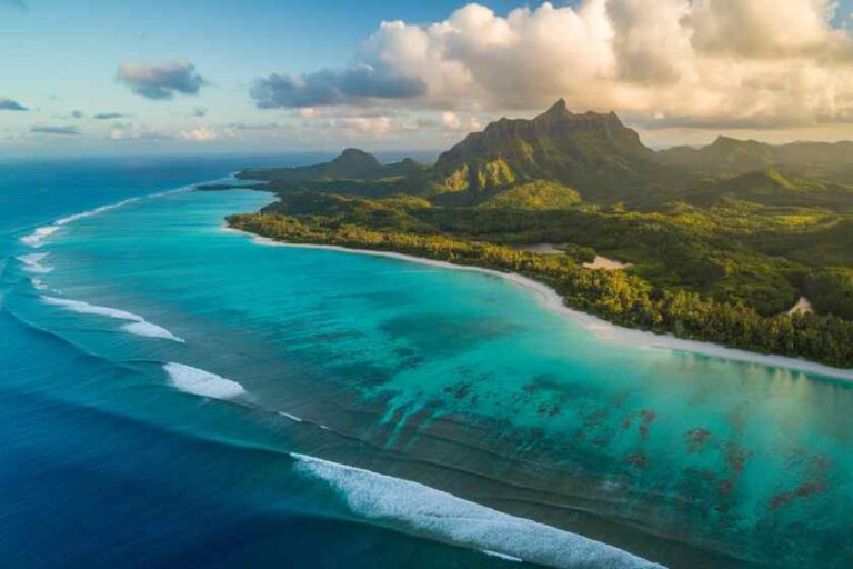 Aerial view of turquoise lagoon along a tropical coastline with lush green mountains in the distance.