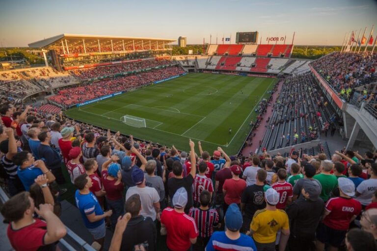 Crowd of soccer fans fills a large stadium, viewing a green pitch with goals as the sun sets behind the stands.