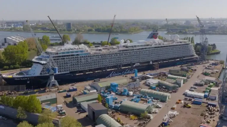 Large cruise ship in a dry dock surrounded by cranes and maintenance tents at a shipyard by the river, Mein Schiff 5 visible on the hull.