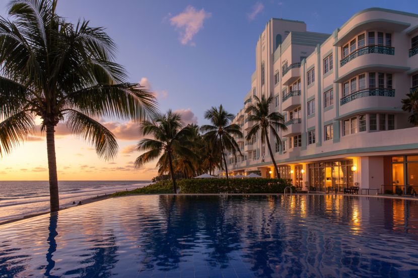 Hotel beside an infinity pool at sunset, palm trees along the waterfront and an ocean view.