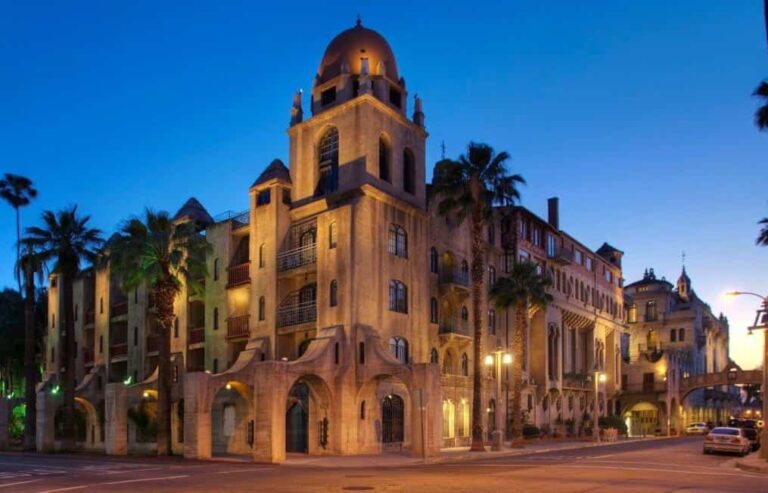 Evening view of a Mediterranean-style building with a domed tower and arched entryway along a palm‑lined street.