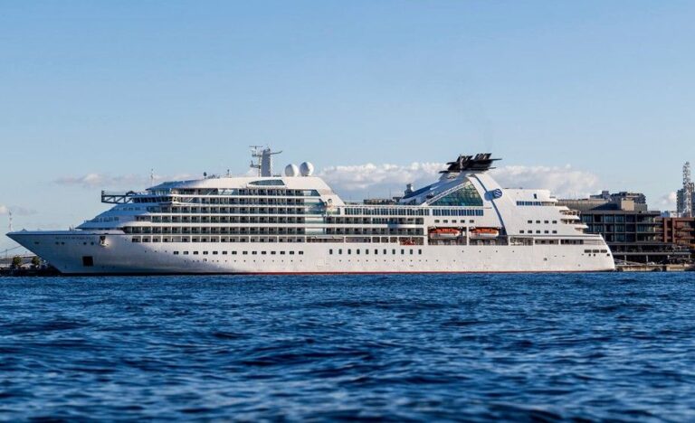Large white cruise ship docked at a harbor, with calm blue water in the foreground and a clear sky above.