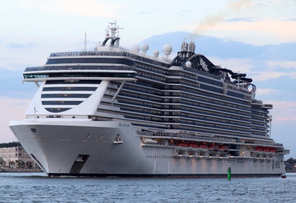 Large white and blue cruise ship docked in a harbor, with multiple decks and lifeboats along its side, calm water in the foreground.