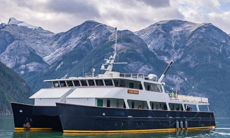 Catamaran-style passenger ferry named Cascadia on calm water with rugged mountain backdrop and cloudy sky.