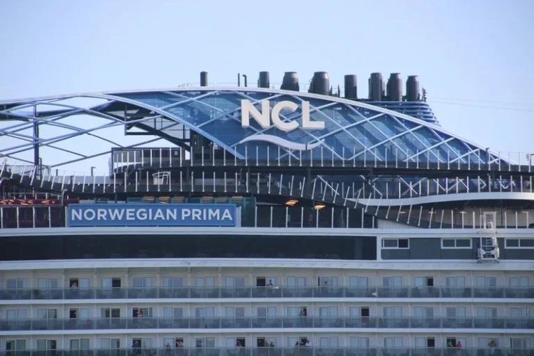 Cruise ship side view featuring the NCL logo and the name 'Norwegian Prima' on the hull, with many balcony cabins visible.