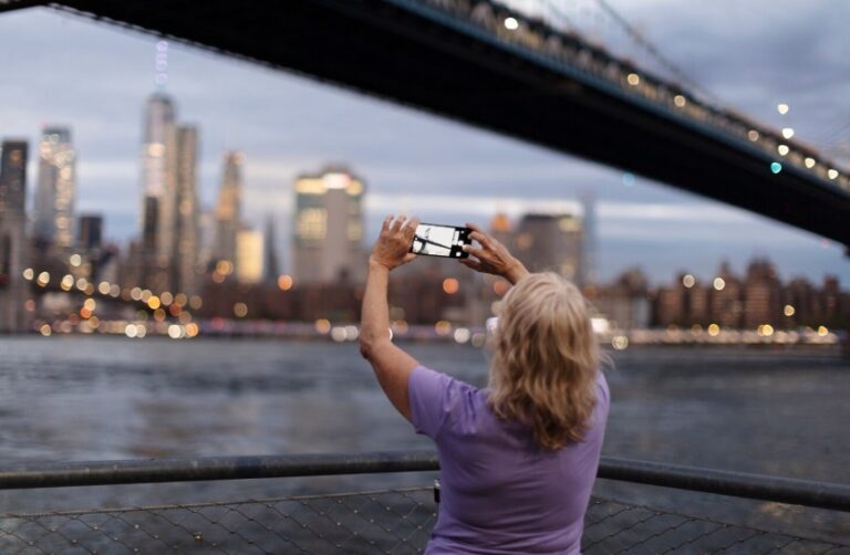 Person in a purple shirt stands at a railing by the water, taking a photo of the illuminated city skyline with a smartphone beneath a large bridge at dusk.