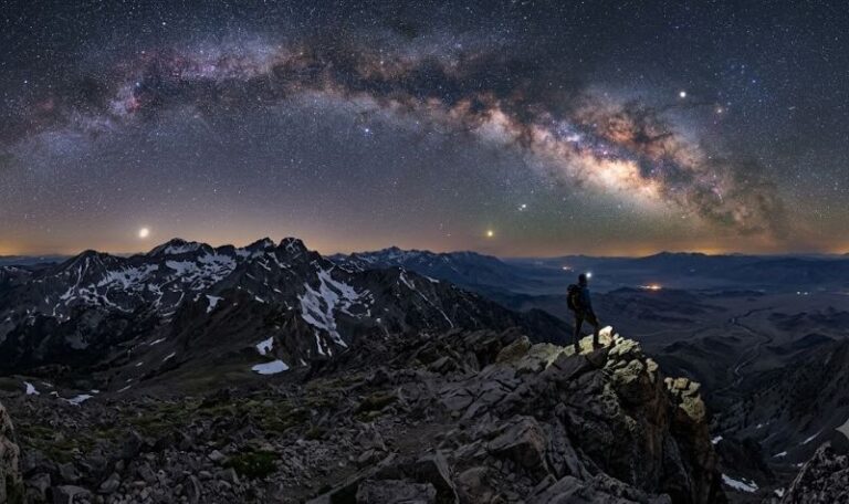 A lone hiker stands on a rocky mountain ridge under a star-filled night sky with the Milky Way arc overhead.
