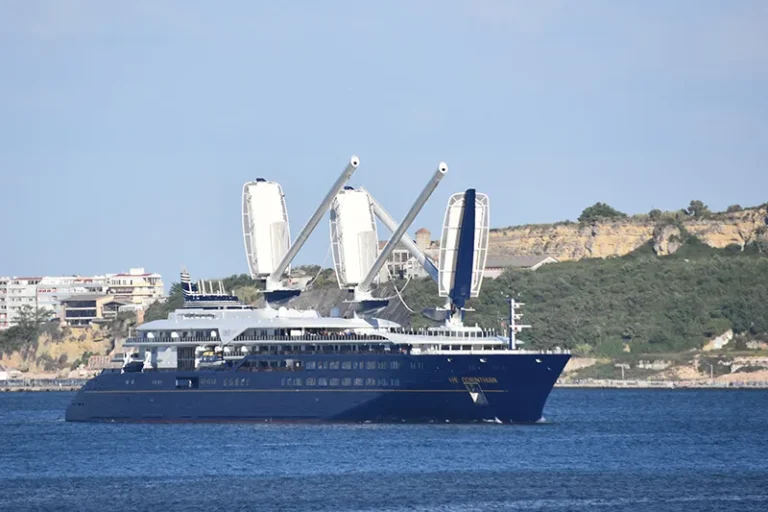 Blue luxury motor yacht anchored near a rocky coastline, with three white crane-like booms on deck.
