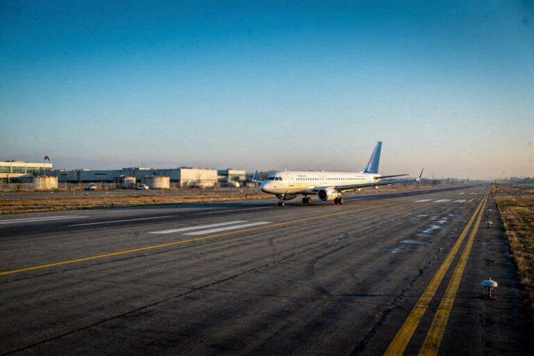 White commercial airplane on a taxiway/runway at an airport during golden hour, heading toward the runway edge.